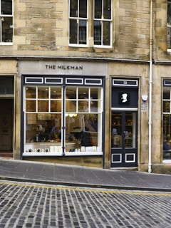A charming café storefront with large windows displaying a cozy interior and various items. A sign on the facade reads 'The Milkman'. The building is made of stone with a traditional design. The street outside is cobblestone, and there is a gentle slope leading up to the cafe's entrance.
