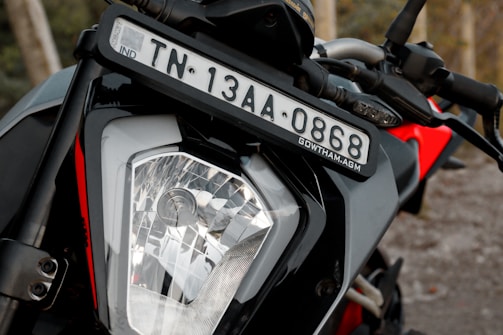 A close-up view of a motorcycle's front section focusing on the headlight and license plate. The motorcycle is parked with handlebars and part of the frame visible. The background is slightly blurred.