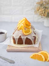 A loaf cake topped with icing and garnished with slices of fresh orange sits on a wooden cutting board. The cake is placed on parchment paper, and a few orange slices are arranged neatly in the foreground. In the background, there is a small white dish and a vase with dried flowers against a white brick wall.