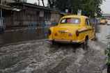 A yellow vintage-style taxi drives through a flooded street, splashing water. The background features roadside stalls and a few other vehicles partially submerged in water.