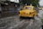 A yellow vintage-style taxi drives through a flooded street, splashing water. The background features roadside stalls and a few other vehicles partially submerged in water.