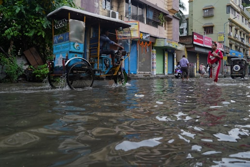 A flooded urban street with waterlogged conditions. People navigate through the water by foot and on bicycles. Shops in the background have closed shutters, and buildings line both sides of the street. A woman in a red outfit is prominently visible walking through the water.