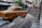 A yellow taxi drives through a flooded street, splashing water as it goes. The surrounding buildings have closed shutters, and several people in the background navigate the floodwaters. The scene appears to be in an urban setting, with signage visible on the buildings.