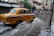 A yellow taxi drives through a flooded street, splashing water as it goes. The surrounding buildings have closed shutters, and several people in the background navigate the floodwaters. The scene appears to be in an urban setting, with signage visible on the buildings.