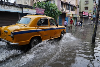 A yellow taxi drives through a flooded street, splashing water as it goes. The surrounding buildings have closed shutters, and several people in the background navigate the floodwaters. The scene appears to be in an urban setting, with signage visible on the buildings.