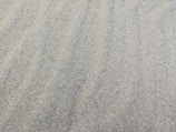 A textured surface of fine, light-colored sand. The sand is evenly spread, creating a smooth and continuous pattern with subtle natural lines and ripples.