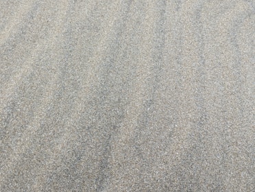 A textured surface of fine, light-colored sand. The sand is evenly spread, creating a smooth and continuous pattern with subtle natural lines and ripples.