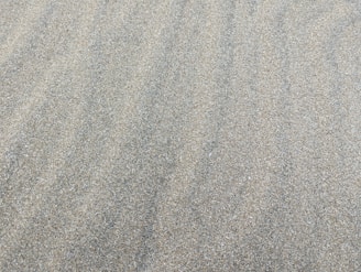 A textured surface of fine, light-colored sand. The sand is evenly spread, creating a smooth and continuous pattern with subtle natural lines and ripples.