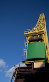 A large industrial crane structure with a distinct yellow lattice framework towers against a clear blue sky. The crane has a green loading container attached, situated prominently with a cautionary sign in Polish below. The framework and machinery details are visible, with some clouds in the sky.