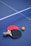 Two table tennis paddles, one with a red face and the other with a black face, are placed next to each other on a blue table. An orange ping pong ball is positioned near the paddles. The net divides the table vertically.