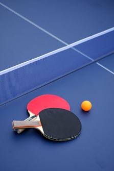 Two table tennis paddles, one with a red face and the other with a black face, are placed next to each other on a blue table. An orange ping pong ball is positioned near the paddles. The net divides the table vertically.