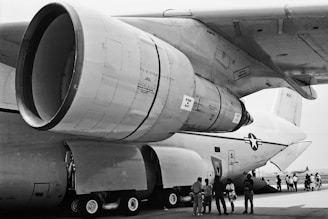 A large military aircraft is parked on an airstrip, with a prominent view of its massive jet engine and landing gear. Several people, including children, are gathered underneath the aircraft, possibly for a tour or demonstration.
