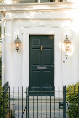 A classic exterior doorway featuring a dark green door with a brass knocker and mail slot, flanked by two wall-mounted lanterns emitting a warm glow. The structure is adorned with white columns and brick, creating a timeless, elegant appearance. A small black iron fence surrounds the entrance, bordered by trimmed hedges. The number 7 is displayed beside the door.