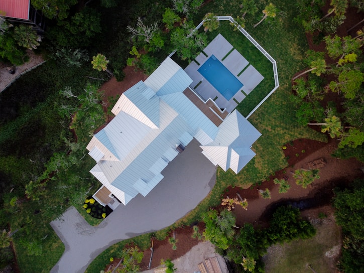 An aerial view of a large house with a light-colored roof surrounded by lush greenery. The property includes a rectangular swimming pool enclosed by a patio with lounge chairs, and a driveway leading to the main building.