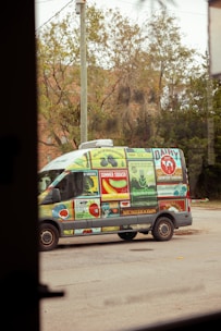 A van is covered in colorful advertisements featuring various fruits, vegetables, and dairy products. The background includes trees and a building partially obscured by foliage.