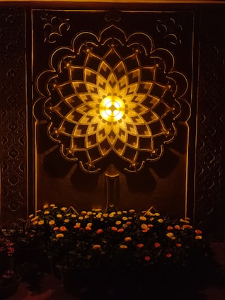 An ornate wall design featuring a geometric pattern with symmetrical shapes radiating from a central light source, casting a warm glow. Below, a bed of flowers with orange and yellow blooms provides contrast.
