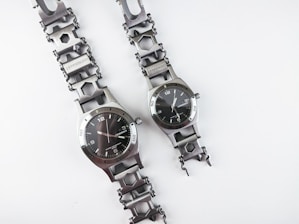 Two metallic wristwatches are placed next to each other on a plain white background. The watches feature unique chain-link bands with hexagonal links and a sleek, industrial design. The watch faces are round with hour, minute, and second hands on a black dial with a minimalist design. Both watches display the brand name prominently.