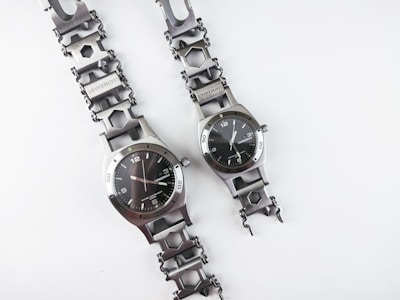 Two metallic wristwatches are placed next to each other on a plain white background. The watches feature unique chain-link bands with hexagonal links and a sleek, industrial design. The watch faces are round with hour, minute, and second hands on a black dial with a minimalist design. Both watches display the brand name prominently.