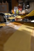 A person is using a manual pasta machine to roll out sheets of fresh pasta dough. The bright kitchen setting features sunlight streaming in, and the scene is focused on the hand guiding the dough through the machine. The workspace is dusted with flour.