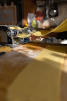 A person is using a manual pasta machine to roll out sheets of fresh pasta dough. The bright kitchen setting features sunlight streaming in, and the scene is focused on the hand guiding the dough through the machine. The workspace is dusted with flour.