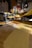 A person is using a manual pasta machine to roll out sheets of fresh pasta dough. The bright kitchen setting features sunlight streaming in, and the scene is focused on the hand guiding the dough through the machine. The workspace is dusted with flour.