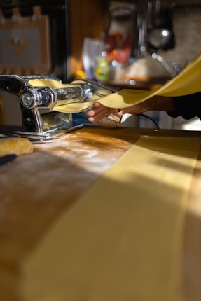 A person is using a manual pasta machine to roll out sheets of fresh pasta dough. The bright kitchen setting features sunlight streaming in, and the scene is focused on the hand guiding the dough through the machine. The workspace is dusted with flour.