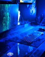 A modern computer setup featuring dual monitors with vivid green and blue graphics. The desk area has a keyboard with illuminated keys and is surrounded by a misty, futuristic blue light. The overall look suggests a high-tech environment.