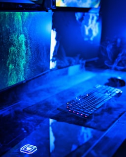 A modern computer setup featuring dual monitors with vivid green and blue graphics. The desk area has a keyboard with illuminated keys and is surrounded by a misty, futuristic blue light. The overall look suggests a high-tech environment.
