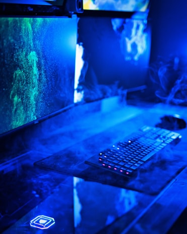 A modern computer setup featuring dual monitors with vivid green and blue graphics. The desk area has a keyboard with illuminated keys and is surrounded by a misty, futuristic blue light. The overall look suggests a high-tech environment.