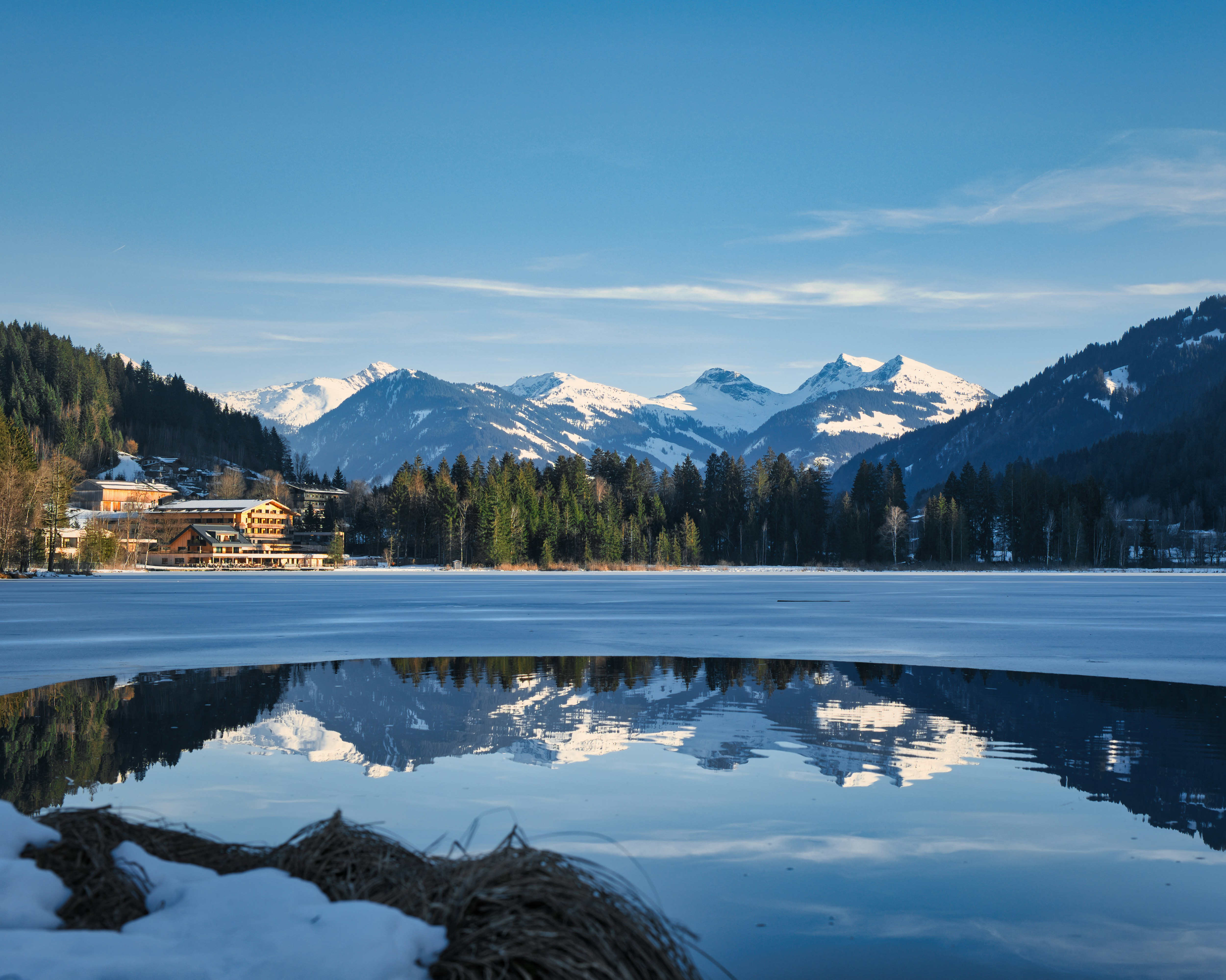 a large body of water surrounded by snow covered mountains, A mountain range reflecting in the schwarzsee at kitzbühel.