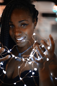 A person with braided hair smiles while holding a string of glowing fairy lights. The lights illuminate their face and hands, creating a warm and intriguing atmosphere.
