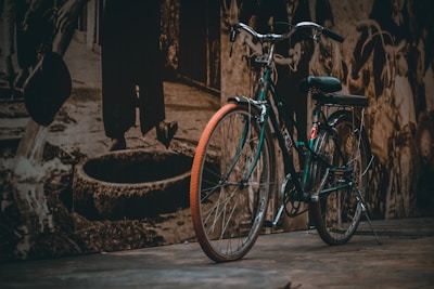 A vintage bicycle with a retro design is parked on a wooden surface, with a sepia-toned mural in the background depicting people in various historical activities. The bicycle features a metal frame and black seat, with distinct details highlighted by contrasting lighting.