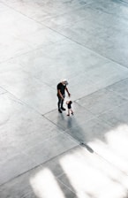A spacious, tiled area with a minimalistic design features two figures: an adult and a child. The adult is wearing a cap and dark clothing, while the child is dressed in light shorts with bright footwear. The open, empty space around them emphasizes their presence and interaction.