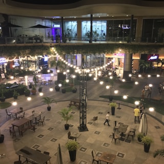 A spacious outdoor courtyard at night with strings of white lights hanging overhead. There are tables and chairs arranged neatly around the area and several potted plants placed throughout. People are walking and sitting within the space, and shops or restaurants with illuminated signs are visible in the background.
