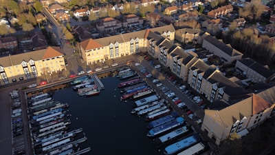Aerial view of a residential area with several apartment buildings surrounding a marina filled with narrowboats. The buildings are arranged in a U-shape, and the marina is enclosed within this formation. Cars are parked along the streets next to the buildings. The area is surrounded by roads and other residential neighborhoods, with some trees interspersed.