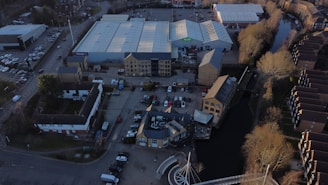 An aerial view of an urban area featuring warehouses with large, white roofs, a parking lot with numerous cars, and several smaller buildings. There is a canal running alongside the buildings, bordered by leafless trees. Residential or office buildings are visible along the canal.