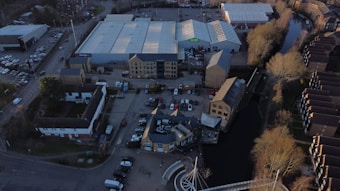 An aerial view of an urban area featuring warehouses with large, white roofs, a parking lot with numerous cars, and several smaller buildings. There is a canal running alongside the buildings, bordered by leafless trees. Residential or office buildings are visible along the canal.