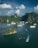 Cruise ships drift peacefully on turquoise waters surrounded by towering limestone cliffs lush with greenery. The contrast of the calm sea with the vibrant greenery and cloudy blue sky creates a picturesque setting.