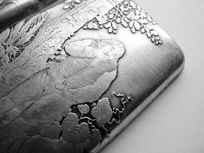 An intricately engraved metal surface depicting a detailed scene of a man with long hair and a vest sitting under a tree. The engraving includes elements like leaves, flowers, and a smoking pipe held by the man.