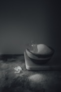 A dimly lit scene featuring a bowl filled with a white granular substance, likely salt, placed on a marble block. A few clear crystal-like objects are scattered nearby, adding an artistic and minimalist touch to the composition.