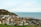 A coastal town with a collection of charming houses with varied architecture nestled against a green hill. The sea stretches into the horizon under a bright, partly cloudy sky. Cars are parked along the seaside, and a small section of the beach is visible.