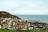 A coastal town with a collection of charming houses with varied architecture nestled against a green hill. The sea stretches into the horizon under a bright, partly cloudy sky. Cars are parked along the seaside, and a small section of the beach is visible.