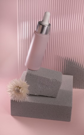 A soft pink and gray composition featuring a cosmetic dropper bottle resting on textured gray blocks. A delicate white flower lies next to the blocks. The background consists of a corrugated pink surface, enhancing the aesthetic appeal with subtle shadows.