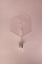 A minimalist arrangement featuring a hexagonal glass or acrylic sheet with several air bubbles encased within. Positioned below is a cosmetic dropper with a metallic cap and a glass body, resting against a soft pink background.