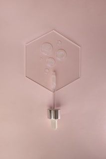 A minimalist arrangement featuring a hexagonal glass or acrylic sheet with several air bubbles encased within. Positioned below is a cosmetic dropper with a metallic cap and a glass body, resting against a soft pink background.