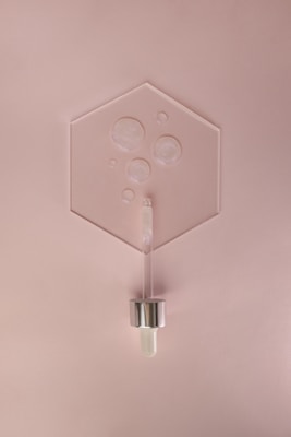 A minimalist arrangement featuring a hexagonal glass or acrylic sheet with several air bubbles encased within. Positioned below is a cosmetic dropper with a metallic cap and a glass body, resting against a soft pink background.