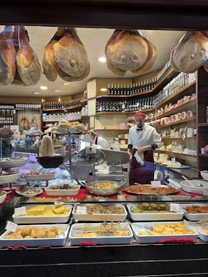 A delicatessen shop interior featuring a wide variety of Italian cured meats and dishes. Large hams are hanging from the ceiling, and numerous trays filled with a selection of prepared delicacies are displayed. Shelves in the background are stocked with bottles of wine and various cheeses. A person wearing an apron and a red cap is working behind the counter, possibly slicing deli meat.