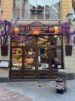 A charming storefront of an Italian shop named 'La Prosciutteria' with rustic wooden doors and a sign displaying various specialties such as 'panini' and 'merende.' The facade is adorned with vibrant purple flowers, adding a touch of elegance. Inside, shelves are filled with food products, creating a warm and inviting atmosphere.