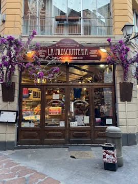 A charming storefront of an Italian shop named 'La Prosciutteria' with rustic wooden doors and a sign displaying various specialties such as 'panini' and 'merende.' The facade is adorned with vibrant purple flowers, adding a touch of elegance. Inside, shelves are filled with food products, creating a warm and inviting atmosphere.
