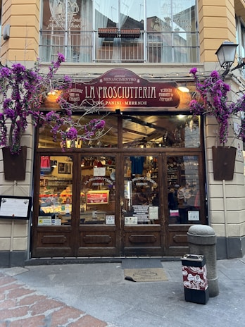 A charming storefront of an Italian shop named 'La Prosciutteria' with rustic wooden doors and a sign displaying various specialties such as 'panini' and 'merende.' The facade is adorned with vibrant purple flowers, adding a touch of elegance. Inside, shelves are filled with food products, creating a warm and inviting atmosphere.